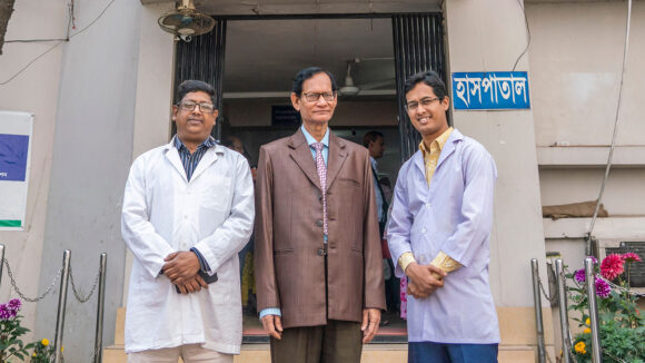 Three men stand outside a hospital entrance.