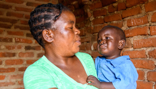 A mother holds and looks at her young son as they stand in front of a brick wall.